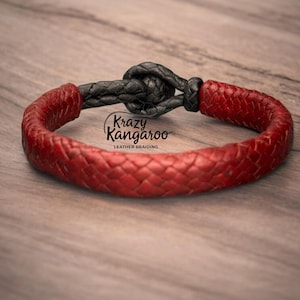May include: A braided red leather bracelet featuring a black leather knot closure. The bracelet displays the text Krazy Kangaroo LEATHER BRAIDING in black lettering. The design consists of a thick, woven red leather band connected by a dark knot.
