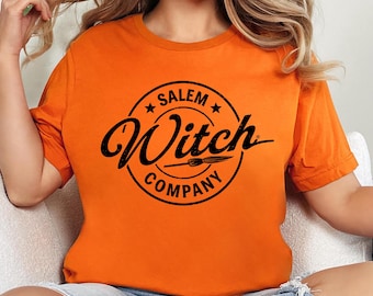 Può includere: T-shirt arancione con grafica circolare nera con la scritta "Salem Witch Company". Il design include stelle e un'illustrazione stilizzata di una forchetta. La maglietta è a maniche corte e in tessuto morbido.