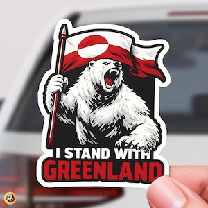 Hand holding a die-cut sticker featuring a roaring polar bear holding the red and white Greenland flag. The text below reads I Stand With Greenland in bold red and white letters on black.