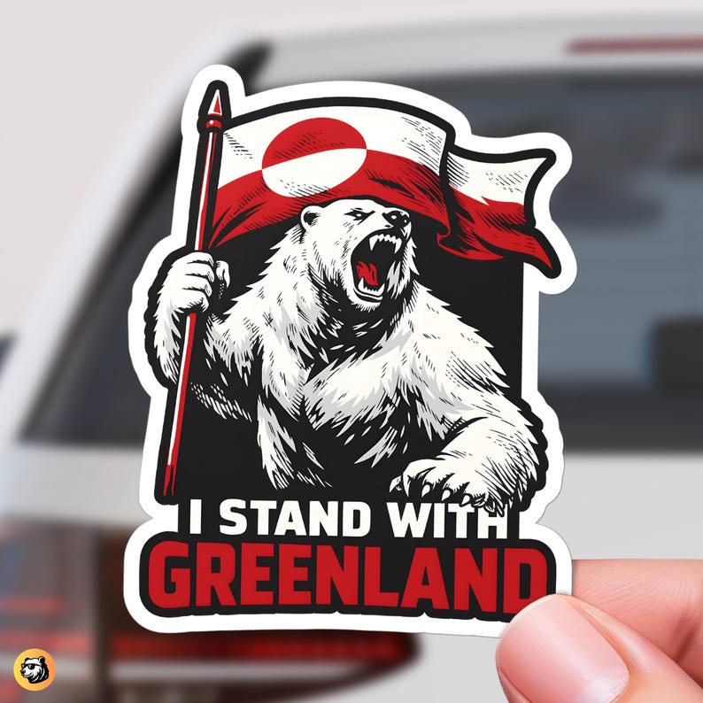 Hand holding a die-cut sticker featuring a roaring polar bear holding the red and white Greenland flag. The text below reads I Stand With Greenland in bold red and white letters on black.