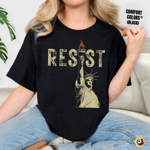 A woman sits comfortably on a cozy sofa, wearing a black Comfort Colors t-shirt with a bold RESIST design. The vintage Lady Liberty print stands out, symbolizing activism and strength. The warm-toned room enhances the relaxed yet empowering vibe of the outfit.