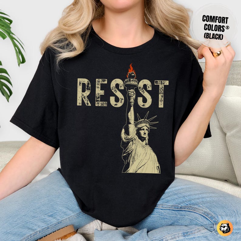 A woman sits comfortably on a cozy sofa, wearing a black Comfort Colors t-shirt with a bold RESIST design. The vintage Lady Liberty print stands out, symbolizing activism and strength. The warm-toned room enhances the relaxed yet empowering vibe of the outfit.