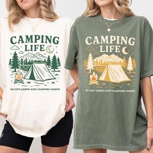 Two women wearing Camping Life Comfort Colors shirts with “No City Lights Just Campfire Nights” graphic; one in ivory, one in moss green. Outdoor-themed vintage camping shirt, perfect for cozy nights, camp trips, and nature lovers.