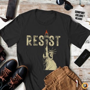 Black Comfort Colors t-shirt featuring a bold RESIST design with a distressed Lady Liberty graphic. The vintage protest print stands out with a fiery torch, symbolizing activism and resistance. Laid flat with stylish accessories, highlighting a casual yet powerful statement piece.