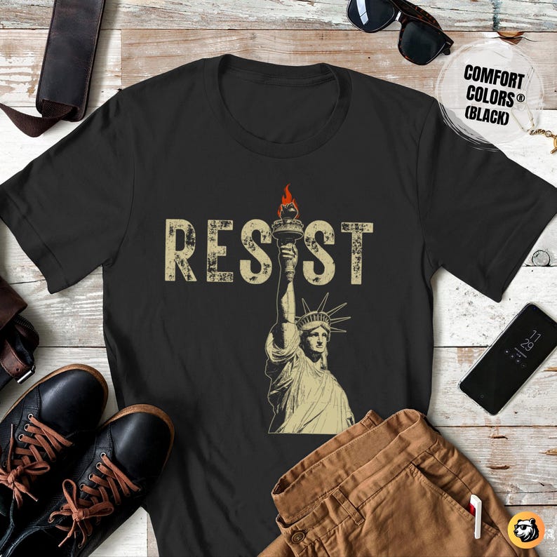 Black Comfort Colors t-shirt featuring a bold RESIST design with a distressed Lady Liberty graphic. The vintage protest print stands out with a fiery torch, symbolizing activism and resistance. Laid flat with stylish accessories, highlighting a casual yet powerful statement piece.