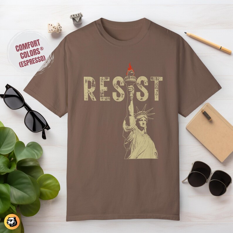 May include: A brown t-shirt featuring a vintage-style image of the Statue of Liberty holding a torch and the word "RESIST" in distressed lettering.