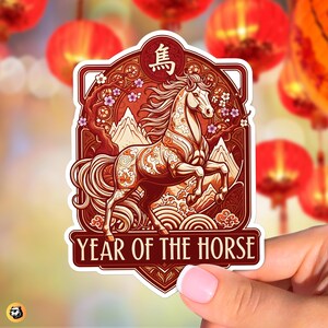 May include: A sticker featuring a prancing horse design with the text "YEAR OF THE HORSE." The sticker has a red and gold colour scheme with floral and mountain accents. The Chinese character for horse is at the top.