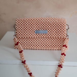 May include: A small pink beaded bag with a silver metal tag that says "ARITELLI" and a pink beaded strap with red beads.