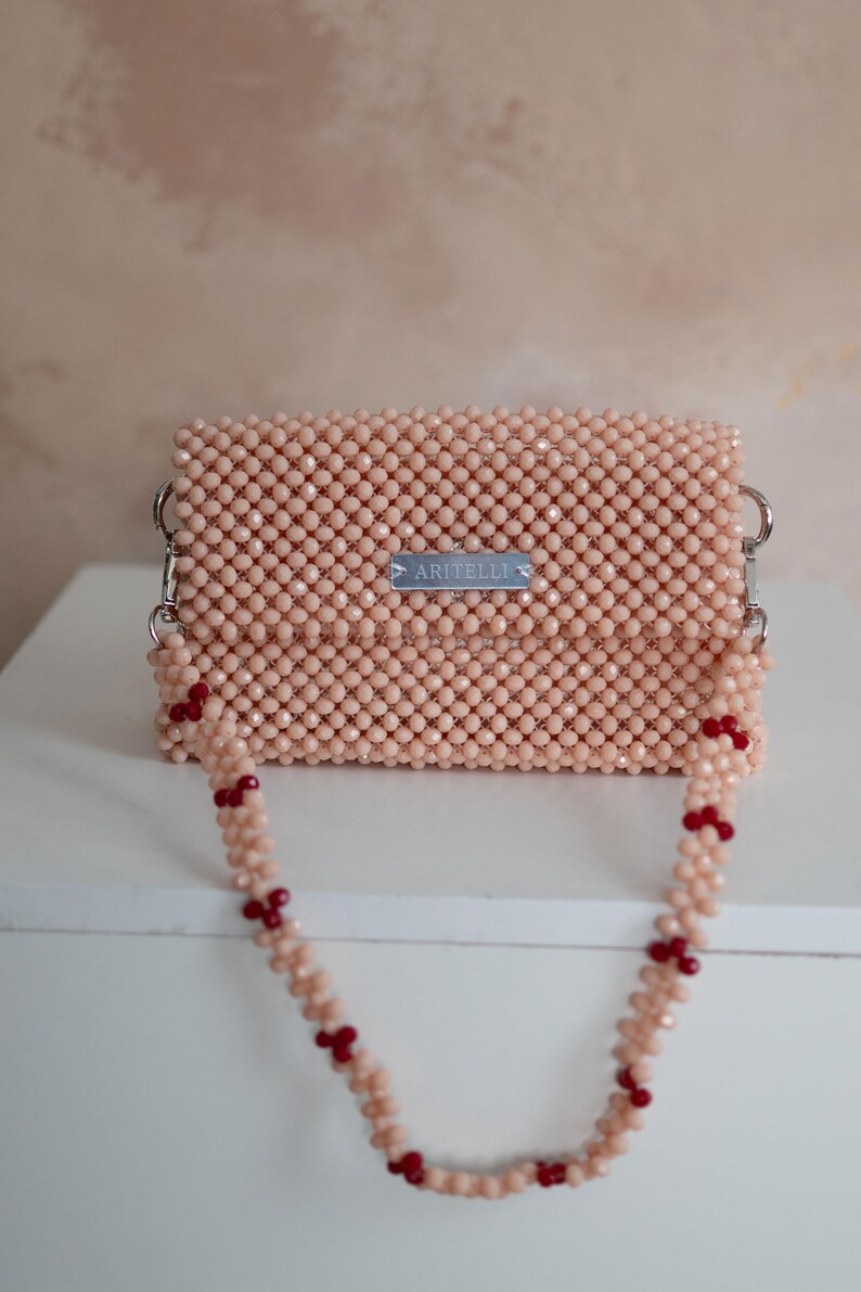 May include: A small pink beaded bag with a silver metal tag that says "ARITELLI" and a pink beaded strap with red beads.