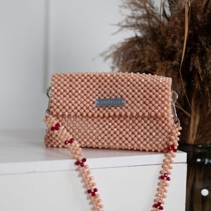 May include: A light pink beaded clutch bag with a matching beaded strap featuring red accents. The bag has a silver label that reads "ARITELLI".
