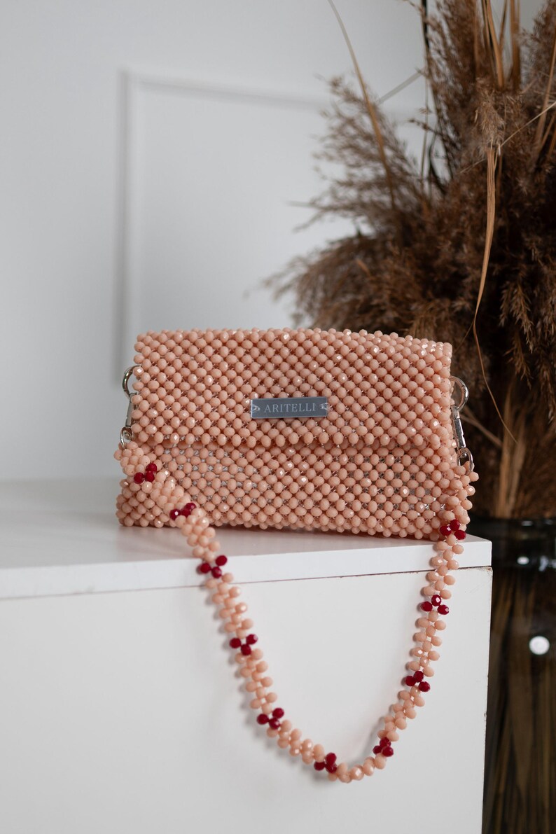 May include: A light pink beaded clutch bag with a matching beaded strap featuring red accents. The bag has a silver label that reads "ARITELLI".