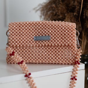May include: A light pink beaded clutch bag with a silver chain strap and red beads. The bag has a small silver label that reads "ARTIELLI."