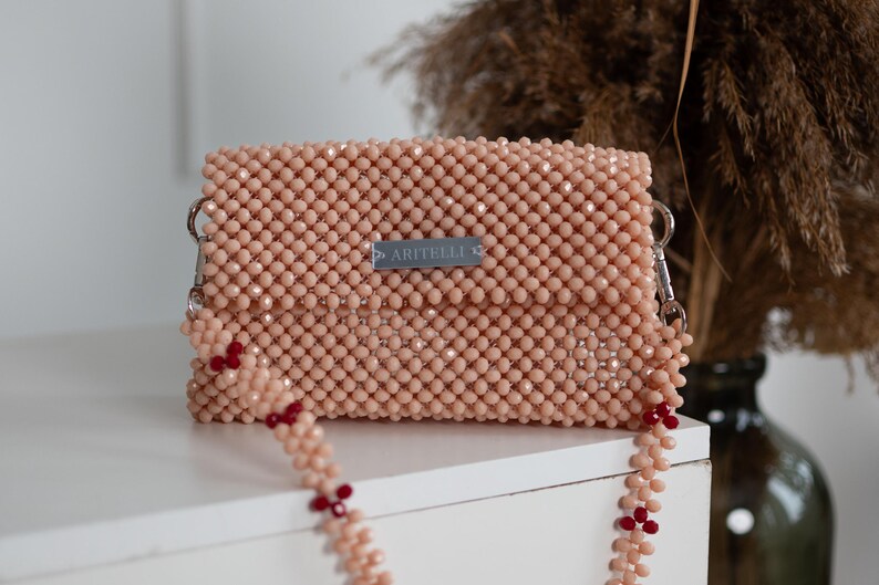 May include: A light pink beaded clutch bag with a silver chain strap and red beads. The bag has a small silver label that reads "ARTIELLI."