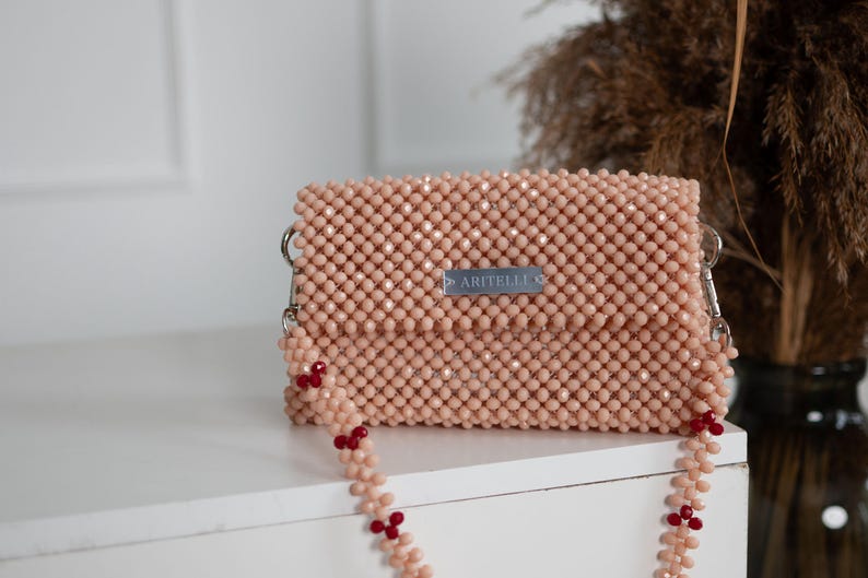 May include: A light pink beaded crossbody bag with a silver metal clasp and a red beaded strap. The bag has a small silver label that reads "ARITELLE C".
