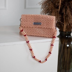 May include: A peach-colored beaded handbag with a red beaded strap. The bag has a silver metal tag with the word "ARITELLI" on it.