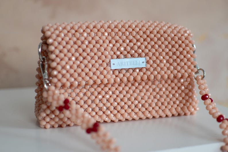 May include: A light pink beaded handbag with a silver clasp and a red beaded strap. The bag has a small white label with the text "ARTITELLI" on it.