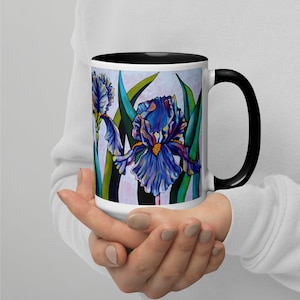 May include: A white ceramic mug with a black rim and handle features a colorful illustration of blue, purple, and green iris flowers. The mug is held in a person's hands.