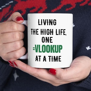 Op de afbeelding: Witte keramische mok met zwarte tekst die "Living the high life, one =VLOOKUP at a time" luidt. De tekst "=VLOOKUP" is groen.