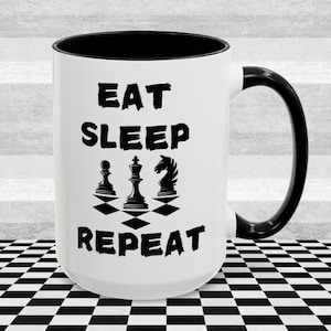 Może przedstawiać: Biały kubek ceramiczny z czarnym rantem i uchwytem. Kubek ma czarny nadruk z napisem "Eat Sleep Repeat" z figurami szachowymi na szachownicy między słowami.