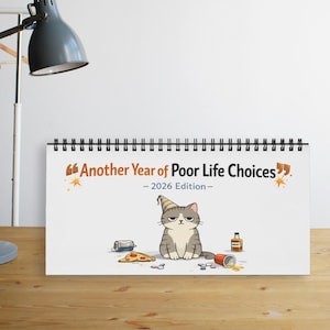 May include: A spiral-bound calendar with the text "Another Year of Poor Life Choices - 2026 Edition." The calendar features a cartoon cat with a party hat, pizza, and spilled drink. A can and a bottle are also visible.