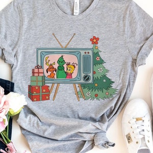 May include: A heather gray t-shirt featuring a vintage television graphic with Christmas characters and a decorated tree. The tee is paired with blue jeans, white sneakers, and a bouquet of pink and white flowers.
