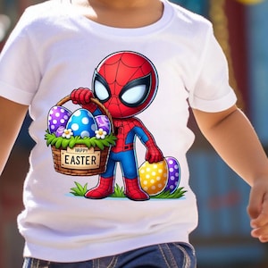 以下が含まれることがあります： カラフルなイースターエッグの入ったバスケットを持ったスパイダーマンの漫画の画像がプリントされた白いTシャツ。バスケットには「ハッピーイースター」の文字が書かれています。若い男の子がシャツを着て笑顔を見せています。
