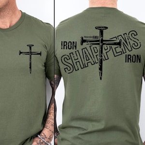 May include: Olive green t-shirt featuring a black nail cross design on the front and back. The back of the shirt has the text "Iron Sharpens Iron" with the Bible verse Proverbs 27:17.