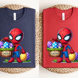 以下が含まれることがあります： イースターエッグの入ったバスケットを持ったスパイダーマンの漫画の画像がプリントされたTシャツが2枚。バスケットには「ハッピーイースター」の文字が書かれている。1枚はネイビーブルー、もう1枚はレッド。