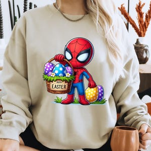 以下が含まれることがあります： イースターエッグの入ったバスケットを持ったスパイダーマンの漫画の画像がプリントされたベージュのトレーナー。バスケットは花と「ハッピーイースター」の文字で飾られています。
