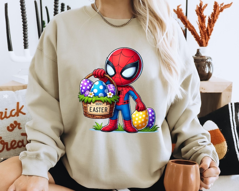 以下が含まれることがあります： イースターエッグの入ったバスケットを持ったスパイダーマンの漫画の画像がプリントされたベージュのトレーナー。バスケットは花と「ハッピーイースター」の文字で飾られています。