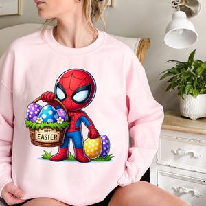 以下が含まれることがあります： ピンクのトレーナーに、イースターエッグの入ったバスケットを持ったスパイダーマンの漫画の画像が描かれています。バスケットには「ハッピーイースター」の文字が書かれています。