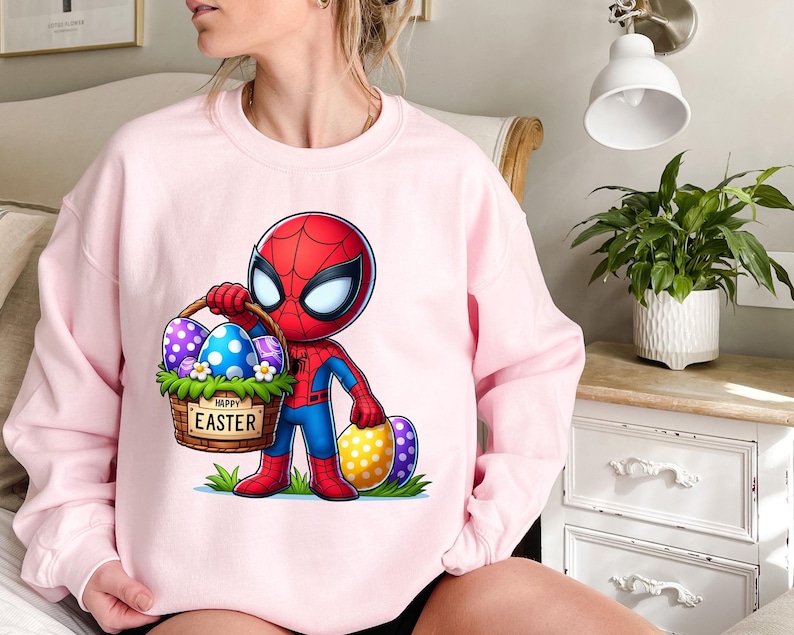 以下が含まれることがあります： ピンクのトレーナーに、イースターエッグの入ったバスケットを持ったスパイダーマンの漫画の画像が描かれています。バスケットには「ハッピーイースター」の文字が書かれています。