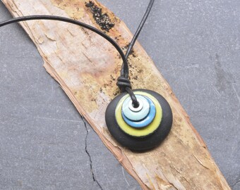 May include: A necklace featuring a black leather cord and a pendant composed of a large matte black circular base topped with three smaller, stacked, concentric rings in yellow, blue, and light blue, finished with a central silver-toned metal eyelet.