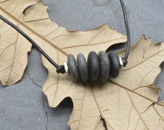 May include: A necklace featuring a dark brown leather cord strung with four smooth, rounded gray stones and two small, square silver-toned metal spacers. The stones are arranged in a row, separated by the metallic accents along the cord.