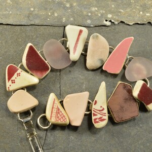 May include: A bracelet made of various pieces of ceramic and glass. The pieces are all different shapes and sizes, and they are connected together with metal rings. The bracelet is a mix of pink, red, and white colors.