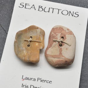 Peut inclure: Deux boutons irréguliers et plats en forme de pierre fixés sur une carte blanche avec le texte SEA BUTTONS, Laura Pierce et Iris Design. Le bouton de gauche est beige avec des veines blanches, celui de droite est rose et beige chiné.