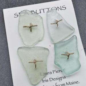 May include: Four sea glass buttons with a cross-shaped design made from twine. The buttons are various shapes and shades of green and clear. The buttons are on a white card with the text "Sea Buttons".