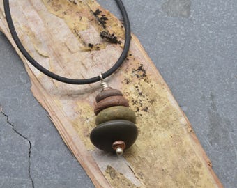 May include: A necklace featuring a black cord and a pendant composed of four stacked, smooth, rounded stones in shades of brown, olive green, and dark gray, accented with small metallic beads at the top and bottom of the stone stack.