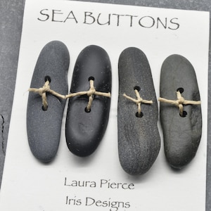 May include: Four elongated, smooth, dark grey and black stone buttons attached to a white display card with tan twine. The card features the text SEA BUTTONS at the top and Laura Pierce Iris Designs printed at the bottom in black ink.