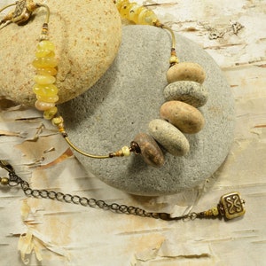 May include: A necklace with a brass chain and a pendant made of a small square with a decorative design. The necklace also features a series of round, natural stone beads in shades of brown and beige.