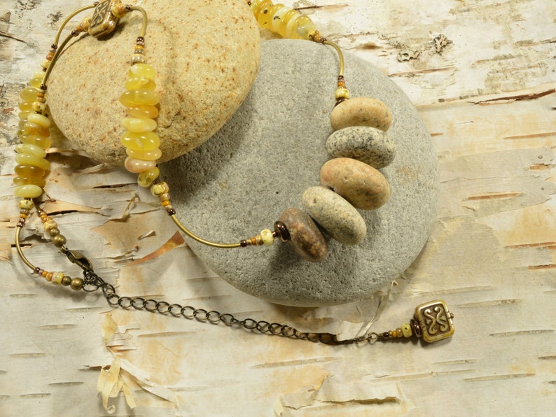 May include: A necklace with a brass chain and a pendant made of a small square with a decorative design. The necklace also features a series of round, natural stone beads in shades of brown and beige.