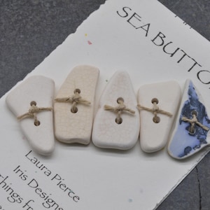 May include: Five sea buttons of varying shapes and colors, including white and a blue and white patterned button. Each button is attached with twine and rests on a white card with the text "SEA BUTTONS".