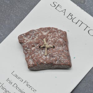 May include: A single, irregular, reddish-brown textured button with a rough, stone-like surface, featuring two central holes threaded with light tan twine. The button is attached to a white display card printed with the text SEA BUTTONS.