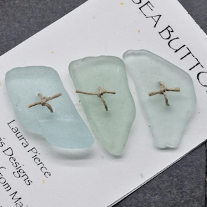 May include: Three sea glass buttons in shades of blue and green, each with a small, cross-shaped twine detail. The buttons are displayed on a white card with the text "SEA BUTTONS" and the maker's name.