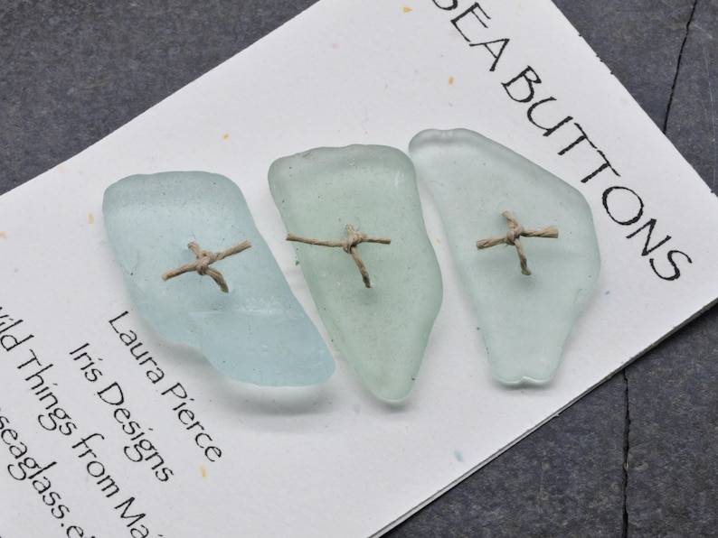 May include: Three sea glass buttons in shades of blue and green, each with a small, cross-shaped twine detail. The buttons are displayed on a white card with the text "SEA BUTTONS" and the maker's name.