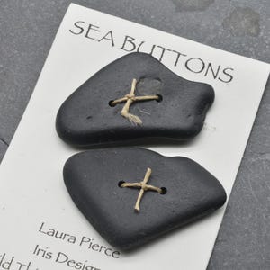 May include: Two dark gray, smooth, irregularly shaped buttons attached to a white display card with light tan twine. The card features the text SEA BUTTONS at the top, with Laura Pierce and Iris Design printed in black text at the bottom.