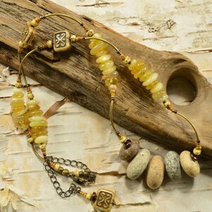 May include: A multi-strand necklace with yellow and brown beads, including smooth, round stones and decorative metal pieces. The necklace is displayed on a piece of driftwood.