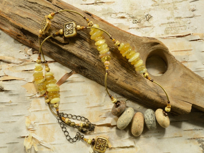May include: A multi-strand necklace with yellow and brown beads, including smooth, round stones and decorative metal pieces. The necklace is displayed on a piece of driftwood.