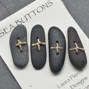 May include: Four dark gray, smooth, elongated stone buttons arranged vertically on a white card labeled SEA BUTTONS. Each button features two drilled holes with a light tan twine threaded through in a cross pattern. The card includes the text Laura Pierce Designs from Maine.