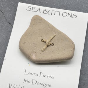Könnte beinhalten: Ein beigefarbener, unregelmäßig geformter Steinknopf, der mit beigefarbenem Garn auf einer weißen Präsentationskarte befestigt ist. Die Karte trägt den Text SEA BUTTONS, Laura Pierce, Iris Designs und Wild. Der Knopf hat eine glatte, gesprenkelte Textur.